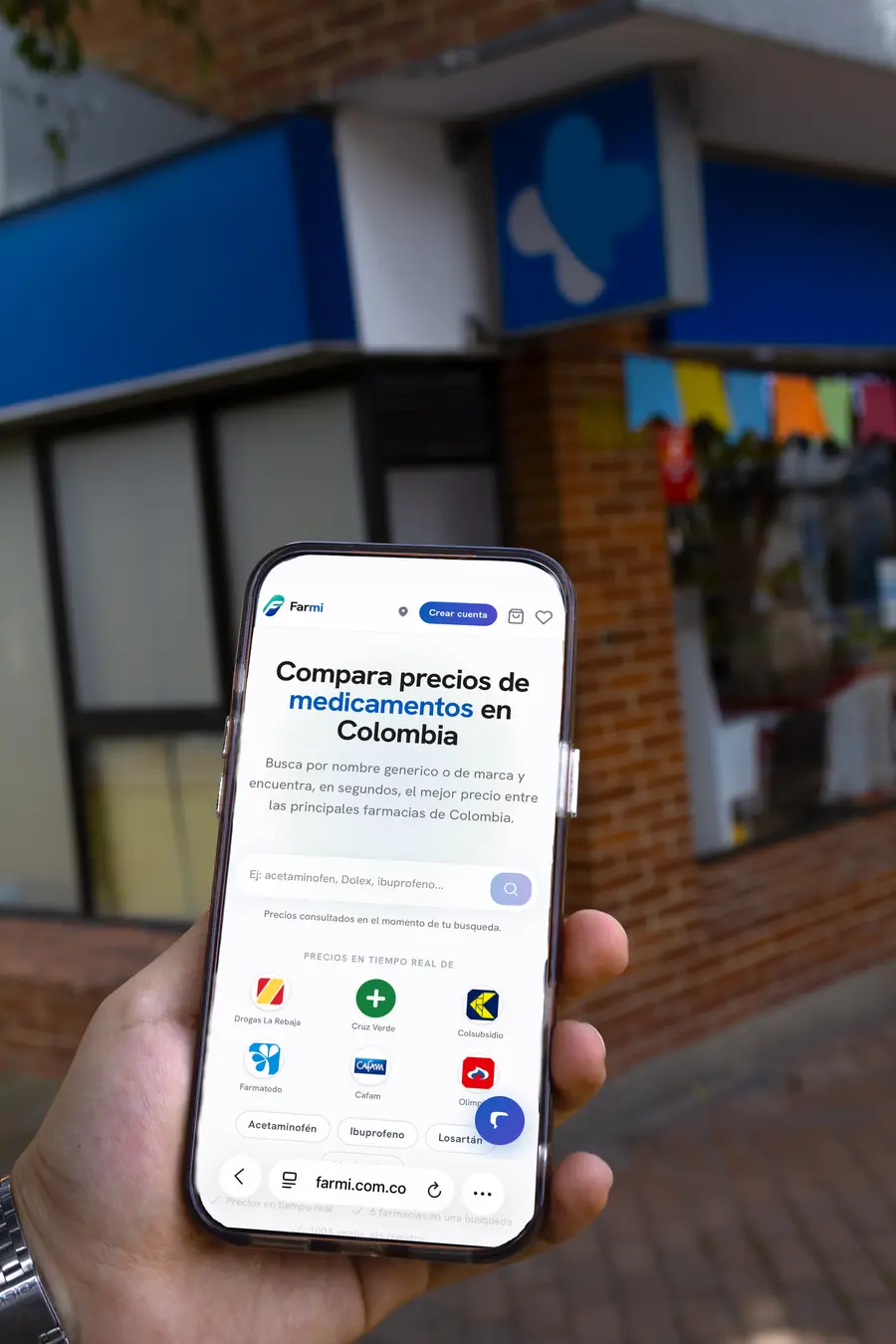 Mano sosteniendo un celular con Farmi abierto frente a una droguería en Colombia
